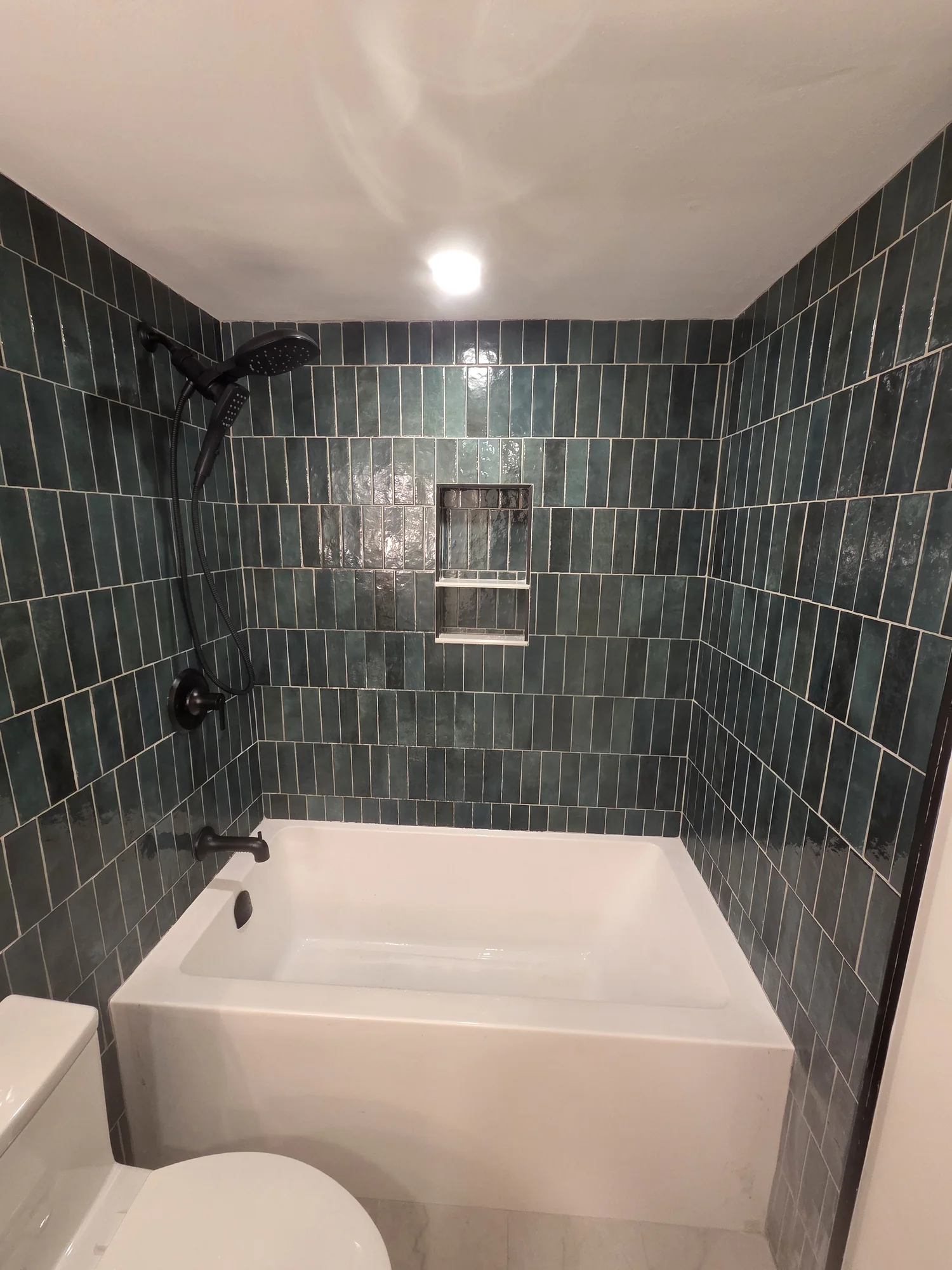 White bathtub and shower in a dark tiled alcove